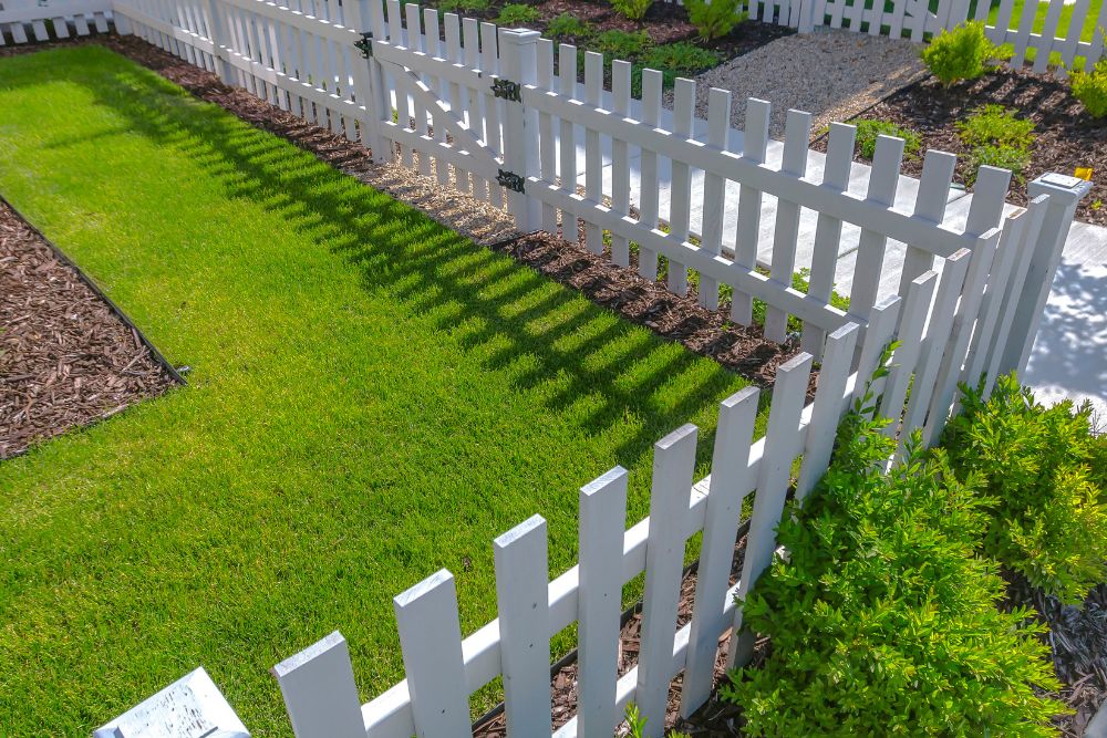A white picket fence surrounds a well-maintained lawn in a sunny yard.